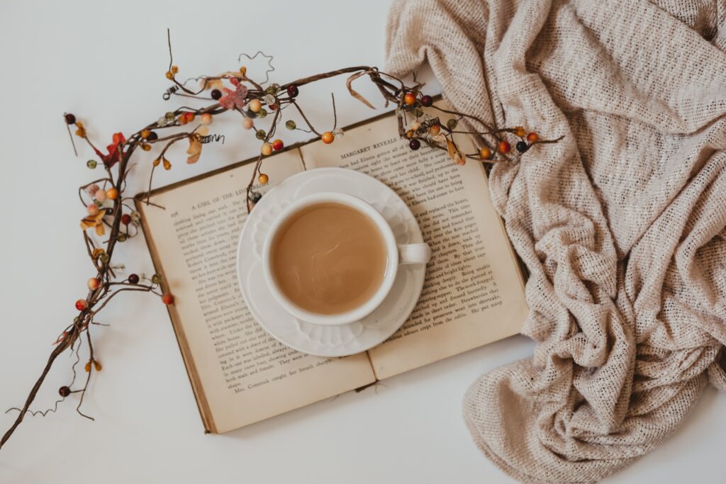 Aware cup of coffee is sitting on top of an open book. Holiday branches and greenery are laid on top of the book. There is a beige blanket bunched around the side of the frame on a white background.