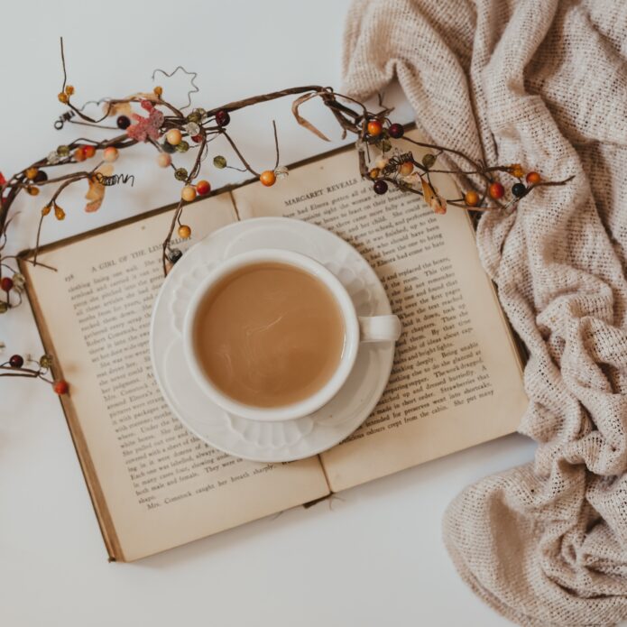 Aware cup of coffee is sitting on top of an open book. Holiday branches and greenery are laid on top of the book. There is a beige blanket bunched around the side of the frame on a white background.