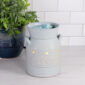 Bring cozy charm to your home with the Blue Blessed Illumination Warmer, designed to look like a vintage milk jug. Featuring a soft matte blue finish with golden flecks, ridged stripe accents, and raised “Blessed” lettering, this ceramic fragrance warmer blends seamlessly into farmhouse, cottagecore, or rustic décor styles.