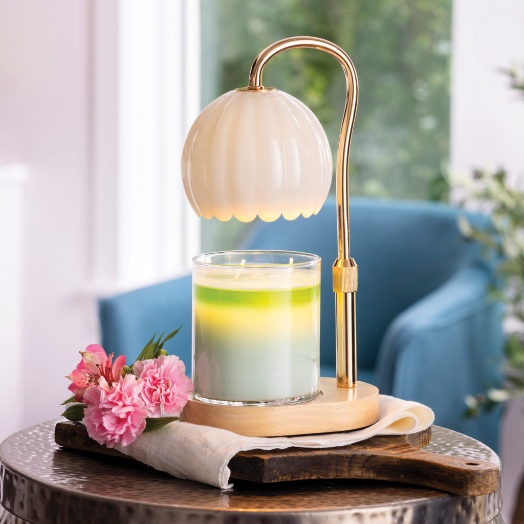 Gold candle warmer lamp illuminating a green layered candle on a side table with pink flowers